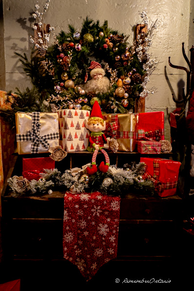 A cute Christmas elf doll sit in front of a pile of Christmas gift boxes on a wooden table.