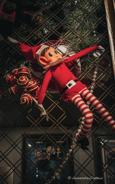 A red Christmas-elf doll hangs from window grilles with a bouquet of ornaments in his hand.