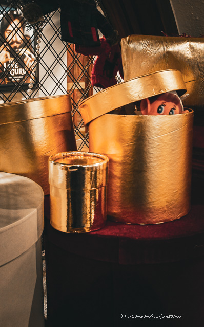 An elf doll peeks from the inside of a gold round present box showing only the eyes.